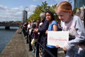 People standing in line, welcoming refugees. Girl holding a banner with text "#RefugeesWelcome".