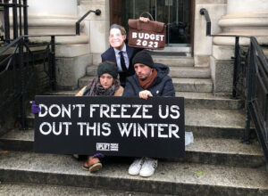 People sitting on stairs outside of the building with poster "Don't freeze us out this winter" 